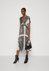 Un vestito midi in blu navy e crema con motivi paisley, maniche corte, design avvolgente, abbinato a stivali alla caviglia neri e a una borsa nera.