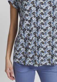 Květinová blůza v světle modré, s černými a žlutými akcenty. Volný střih s ohrnutými rukávy, spárovaná se světle modrými kalhotami, hladká textura.