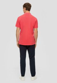 Męska koszulka polo z krótkim rękawem w kolorze koralowym z kontrastowym białym kołnierzykiem, połączona z ciemnoniebieskimi spodniami i białymi trampkami.