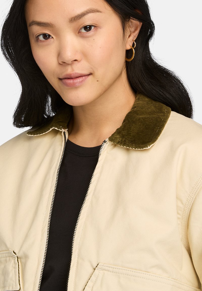 Giacca beige con chiusura a zip e colletto in corduroy verde. Presenta due tasche frontali e una camicia interna nera. Texture liscia.
