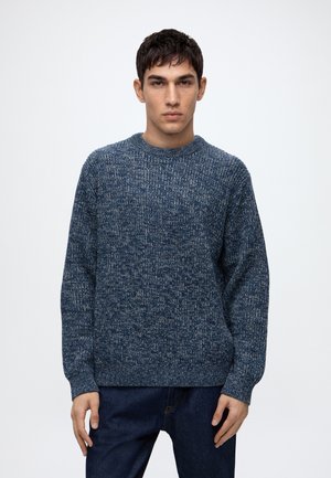 Jeune homme portant un pull en tricot bleu et un jean foncé, debout devant un fond clair uni, face à l'avant.