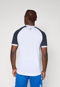 Tricou sport cu mânecă scurtă, cu un corp alb, mâneci raglan negre și un guler cu model. Pe spate se află logo-ul.
