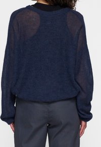 Granatowy dzianinowy sweter o luźnym kroju, przejrzystej teksturze, opuszczonych ramionach i subtelnym logo z tyłu. W zestawie z szarymi spodniami.