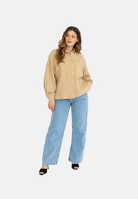 Beżowa koszula z guzikami, wykonana z teksturowanej tkaniny, z kieszonką i luźnym krojem, w połączeniu z jasnoniebieskimi spodniami o szerokich nogawkach i czarnymi szpilkami.