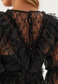 Svart spetsklänning med volangdetaljer, djup V-ringning bak och knappstängning. Intrikat spetsmönster synligt genom hela tyget.