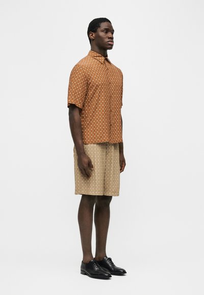 Άνδρας στέκεται με κατεύθυνση προς τα δεξιά, φορώντας πορτοκαλί πουκάμισο με κοντά μανίκια και σχέδια, μπεζ σορτς με σχέδια και μαύρα επίσημα παπούτσια σε λευκό φόντο.