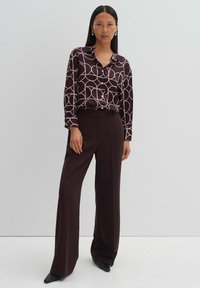 Brown patterned blouse with large white circles, featuring a collar and buttons, paired with high-waisted, dark brown trousers and pointed shoes.