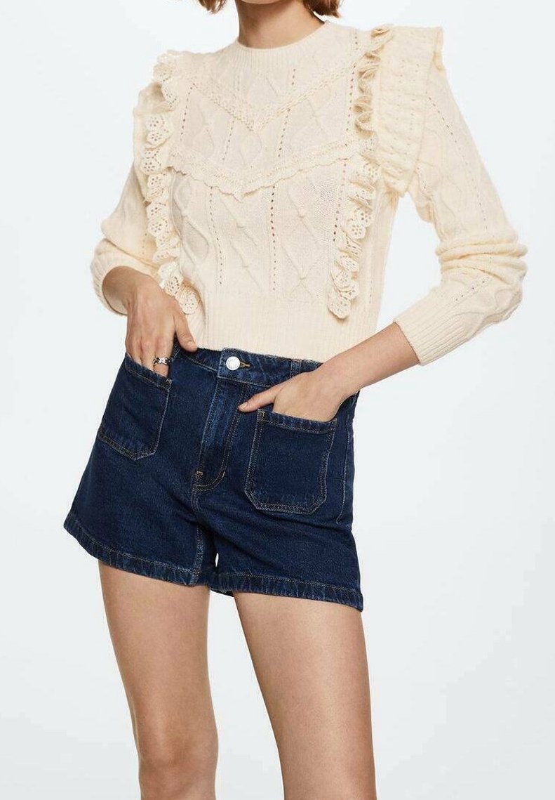 Cremfarbener Strickpullover mit Rüschendetails und strukturierten Mustern, kombiniert mit hochgeschnittenen, dunklen Denim-Shorts mit Fronttaschen.