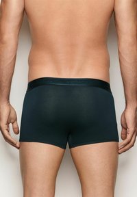Hombre vestido con calzoncillos ajustados negros, de pie contra un fondo claro y liso, visto desde atrás.