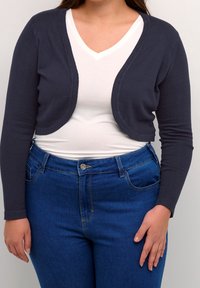 Dunkelblaues, kurzes Strickjacke mit langen Ärmeln, aus weichem Material und abgerundeten Kanten, über einem weißen V-Ausschnitt-Shirt und blauen Jeans kombiniert.
