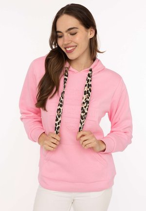 Junge Frau lächelt und hält die leopardbedruckten Kordeln ihres pinken Kapuzenpullovers, trägt weiße Hose und steht vor weißem Hintergrund.
