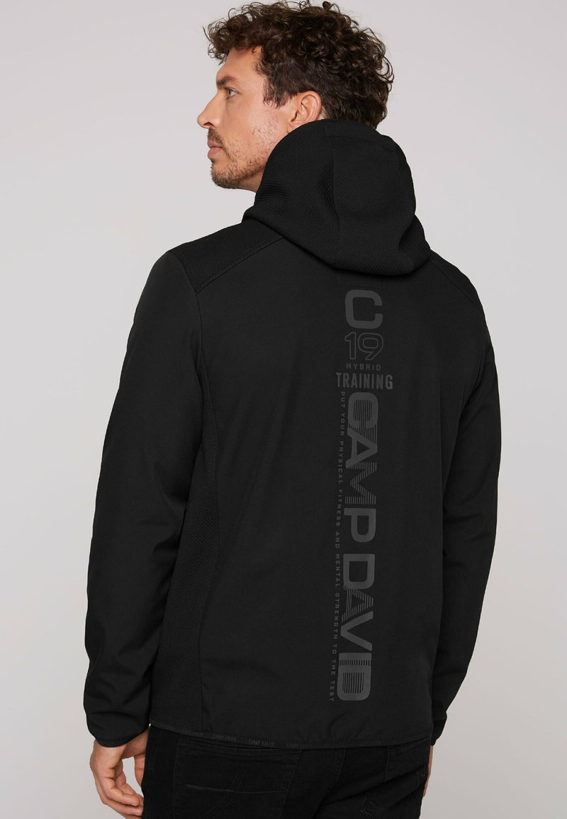 Mustavärinen huppari, joka on valmistettu tekstuurikankaasta ja jossa on pystysuuntainen logopainatus. Muotoilu on vartalonmyötäinen ja pinnassa on sileä viimeistely.