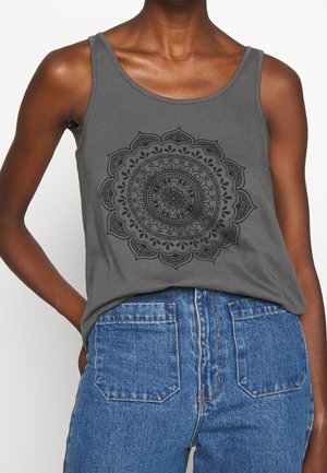 Person wearing sleeveless gray tank top with black intricate circular mandala design and high-waisted blue denim jeans.
