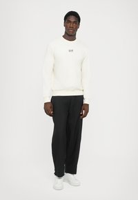 Sudadera blanca de cuello redondo con el logo "EA7", combinada con pantalones de pierna ancha negros y zapatillas blancas, de pie frente a un fondo liso.
