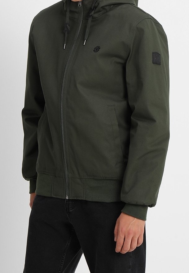 Homme portant une veste zippée vert foncé avec capuche et pantalon noir, debout, la main droite détendue le long du corps.