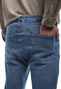 Ciemnoniebieskie dżinsy o dopasowanym kroju, z dwoma tylnymi kieszeniami i widocznymi detalami przeszyć. W połączeniu z beżową bluzką.