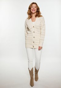 Cardigan bej tricotat cu un model texturat, având un decolteu în V, nasturi pe față și asortat cu pantaloni albaștri și cizme de gleznă maro.