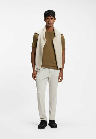 Homme portant un pantalon beige, une chemise marron à manches courtes, un pull crème sur les épaules et des chaussures noires, debout sur un fond blanc.