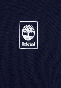 Tejido de punto azul marino con un logotipo rectangular blanco que presenta un diseño de árbol y la palabra "Timberland" debajo.