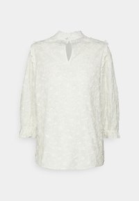 Blouse blanche brodée à manches longues, poignets à volants, petite ouverture en forme de goutte dans le dos, avec une texture florale sur tout le tissu.