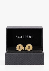 Gemelos de color dorado con un diseño de calavera y huesos cruzados, presentados en una caja negra con un exterior texturizado y un interior de terciopelo.