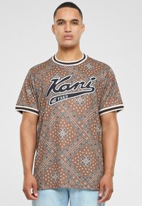 Brūna īsu piedurkņu T-krekls ar paisley rakstu, melnu "Kani" logotipu un baltu svītrainu apdari apkaklē un piedurknēs.