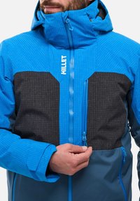 Blaue Outdoorjacke mit Kapuze, ausgestattet mit schwarzen Panelakzenten, Reißverschlusstaschen und einem Karomuster. Hergestellt aus robustem, wetterfestem Material.