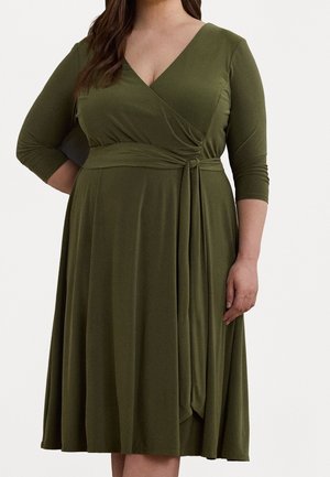 Kvinde iført olivengrøn slå-om-kjole med 3/4-ærmer og bundet taljebånd, stående foran en ensfarvet baggrund.