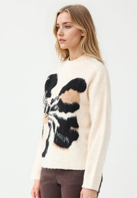 Cremfarbener Pullover mit einem großen schwarzen und tanfarbenen Schmetterlingsdesign, weichem Textilgewebe, Rundhalsausschnitt und langen Ärmeln, kombiniert mit braunen Hosen.