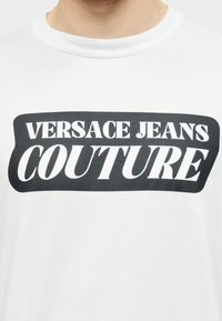 Λευκό κοντομάνικο πουκάμισο με στρογγυλή λαιμόκοψη και μαύρο ορθογώνιο λογότυπο που αναγράφει «Versace Jeans Couture» με έντονα λευκά γράμματα στο στήθος.