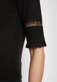 Top de punto negro con mangas cortas con bordes ondulados y detalles de encaje transparente. Textura suave con silueta ceñida.