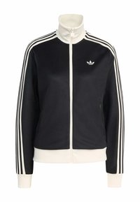 Μαύρο τζάκετ με φερμουάρ και ψηλό γιακά, λευκές διακριτικές ρίγες στα μανίκια και μικρό λογότυπο στο στήθος. Διαθέτει πλαϊνές τσέπες και ριμπ μανσέτες.