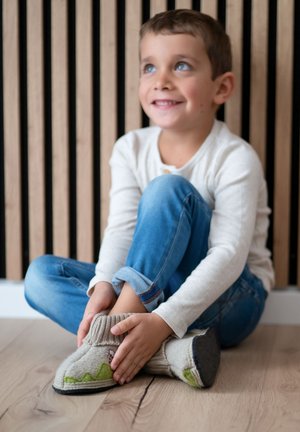 Lächendes Kind sitzt auf dem Boden, trägt ein weißes Langarmshirt, blaue Jeans und richtet beige Wollhausschuhe mit grünen Details.