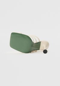 Sandale à talons hauts avec une tige beige, une semelle en caoutchouc verte et un talon fin noir. Présente des fines lanières sur le dessus pour un design minimaliste.