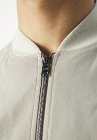 Světle béžová bunda se zipem vpředu. Měkká látka, žebrovaný límec a jemná textura viditelná v látce. Černý kovový zip.