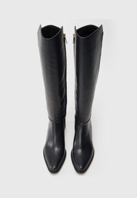 De hautes bottes en cuir noir avec des pointes effilées et des fermetures éclair sur les côtés, droites sur un fond blanc.