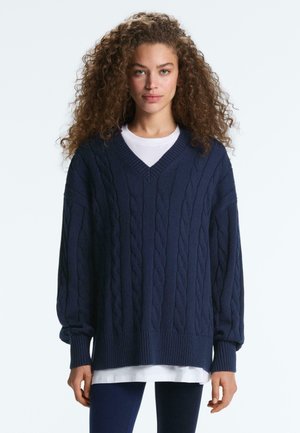 Femme aux cheveux bouclés portant un pull à col en V en maille torsadée bleu marine par-dessus une chemise blanche et un pantalon foncé, debout devant un fond clair.