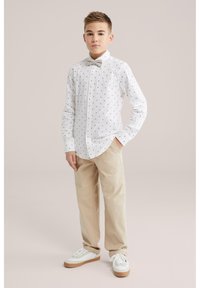 Fiú áll, egyik kezét a zsebében tartja, fehér mintás gombos ingben, bézs nadrágban, bézs csokornyakkendővel és fehér tornacipőben.