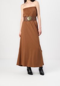 Brązowa maxi sukienka bez ramiączek, z szerokim brązowym paskiem ozdobionym okrągłymi detalami. Czarne botki za kostkę dopełniają stylizację.