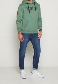 Grön huvtröja med en framficka i känguru-modell och logotyp. Parats med mörkblå jeans och vita sneakers med gröna inslag. Strukturerad tyg.