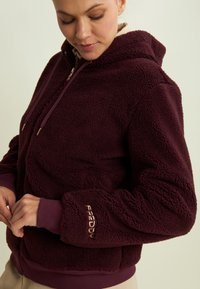 Felpa con zip bordeaux realizzata in pile morbido e texturizzato. Presenta un cappuccio, tasche frontali e un logo ricamato sul polsino.