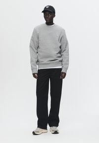 Gray sweatshirt with ribbed cuffs, worn over a white shirt. Black pants and beige sneakers complete the outfit, with a black cap on the head.