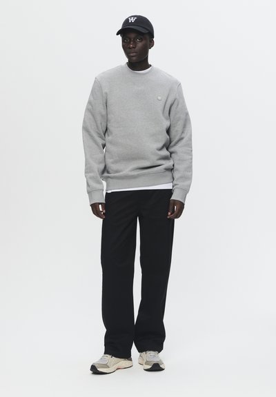 Grå sweatshirt med ribbede manchetter, båret over en hvid skjorte. Sorte bukser og beige sneakers fuldender outfittet, med en sort kasket på hovedet.