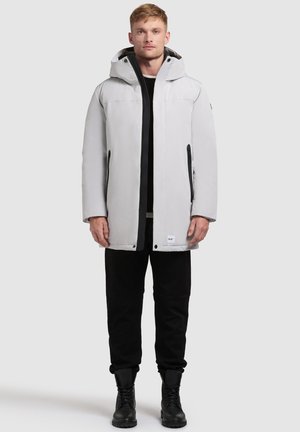Lichtgrijze waterdichte jas met een capuchon, twee zijzakken met rits en een ritssluiting aan de voorkant. Draagt over een zwart shirt en zwarte broek.