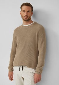 Beige Rippenpullover mit Rundhalsausschnitt und langen Ärmeln. Über einem weißen T-Shirt getragen, kombiniert mit weißen Hosen, die über ein Zugband verfügen.