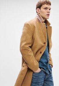 Νεαρός άνδρας με καμηλό παλτό από μαλλί, καρφωμένο σε τζιν πουκάμισο και τζιν παντελόνι, στέκεται με τα χέρια στις τσέπες του μπροστά σε μια απλή λευκή βάση.