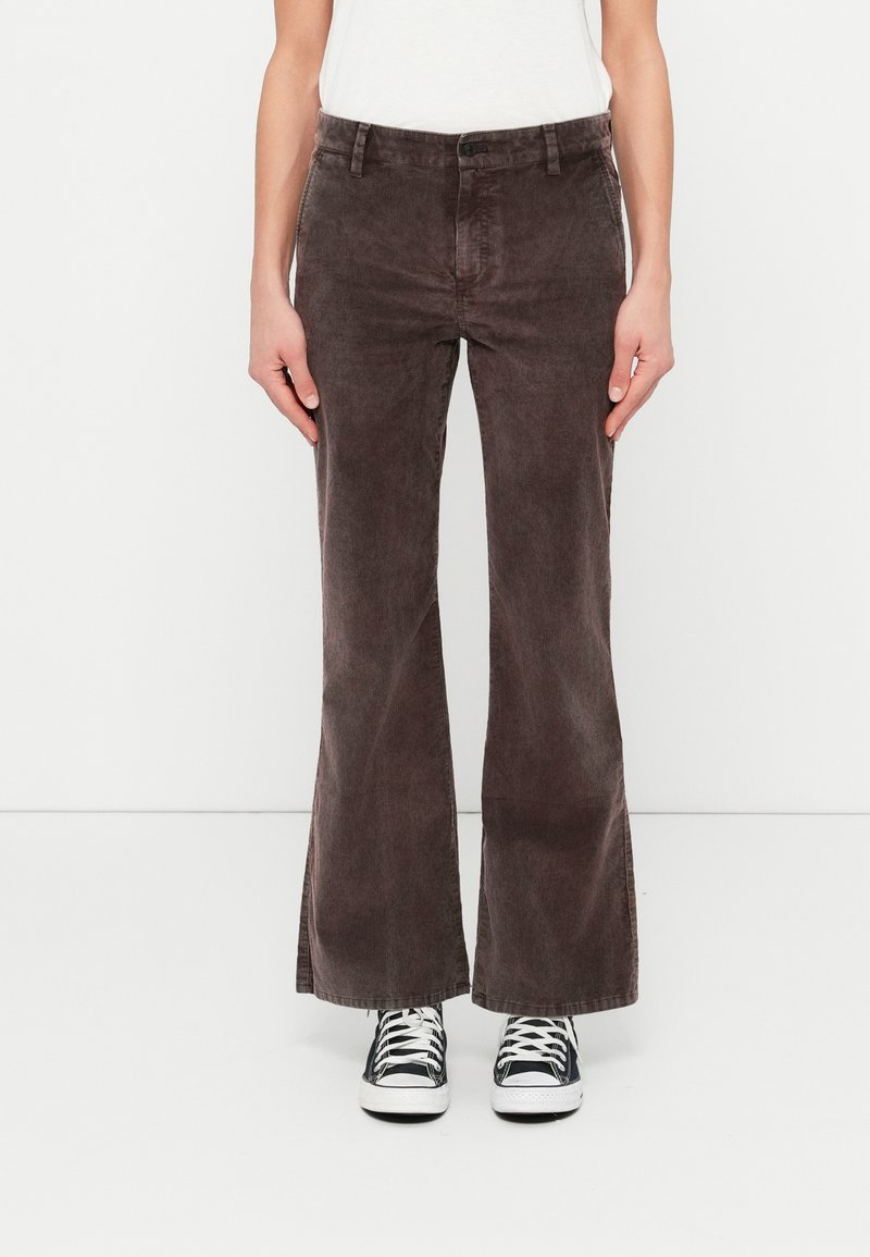 Personne portant un pantalon pattes d'éléphant en velours côtelé marron et des baskets noir et blanc, debout devant un fond blanc uni.