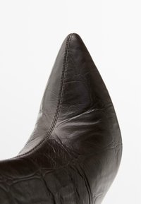 Tmavě hnědá kožená bota s špičatou špičkou, s texturovaným povrchem a viditelnými švy, která předvádí elegantní siluetu.