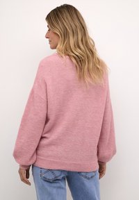 Rózsaszín kötött pulóver laza szabással, süllyesztett vállakkal és bordázott szegéllyel. Világoskék farmernadrággal párosítva. Puha tapintású és oversized dizájnú.