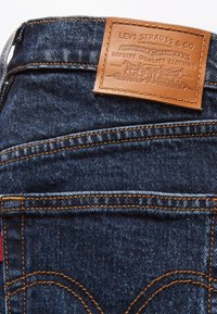 Poche arrière d'un jean en denim bleu foncé avec des surpiqûres orange et un patch en cuir beige Levi Strauss & Co. au-dessus de la poche.